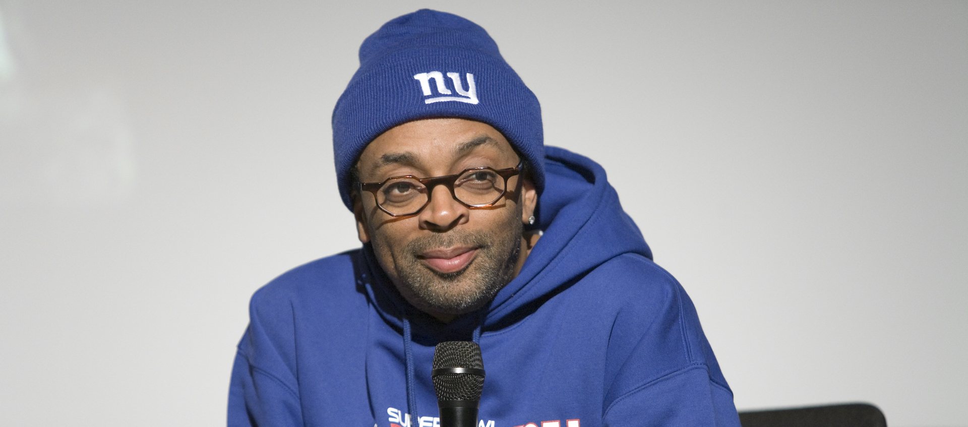 Spike Lee sits in front of a projection screen in a blue New York Knicks sweatshirt and matching knit cap