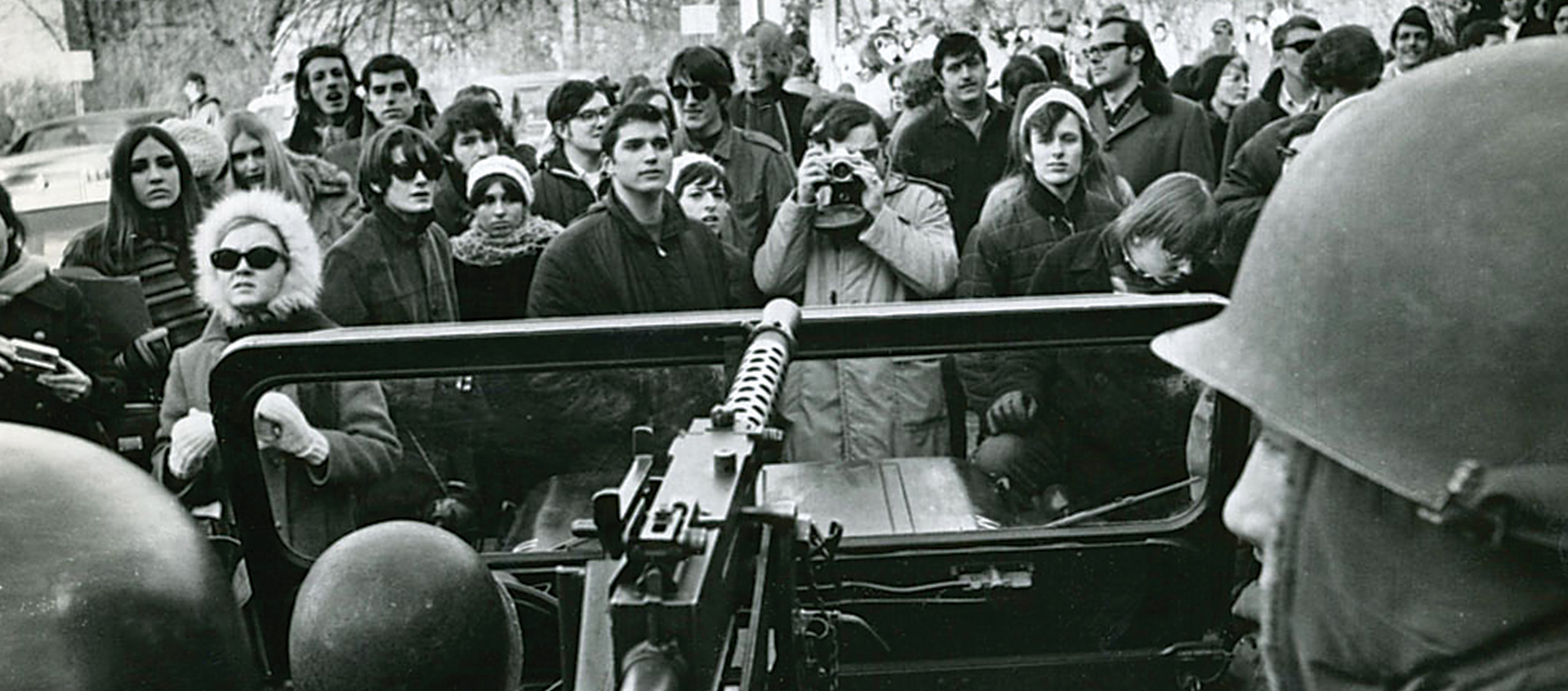 A crowd of protestors stand in front of a jeep carrying soldiers
