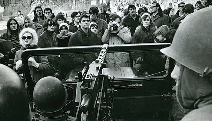 A crowd of protestors stand in front of a jeep carrying soldiers