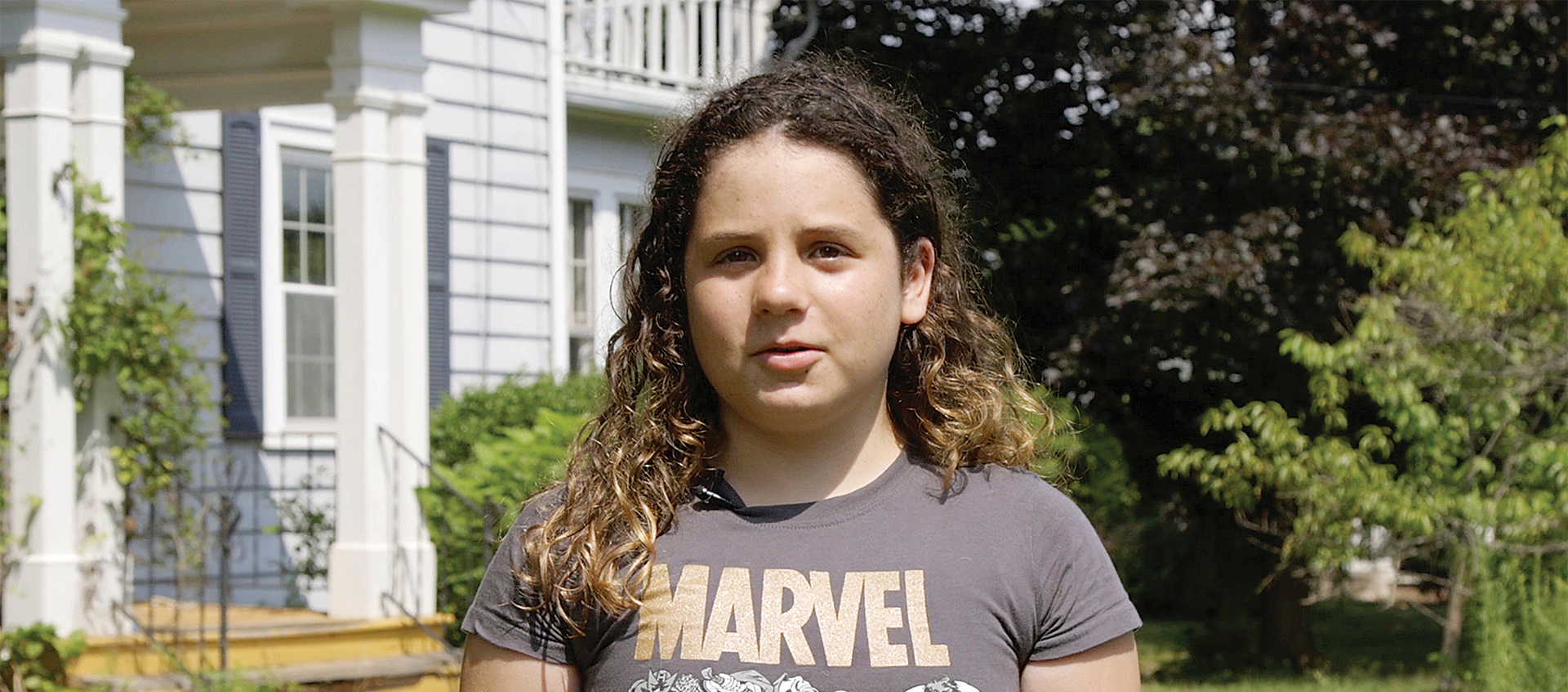 A girl in a Marvel T-shirt stands in front of a home