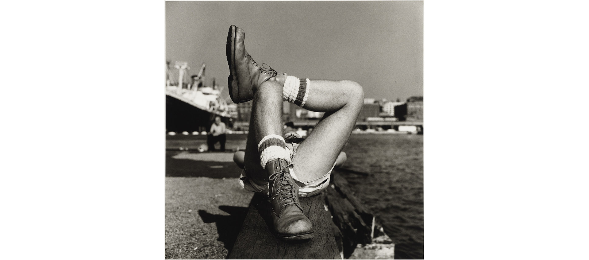 A 1976 Peter Hujar photograph of a man's legs in shorts and boots, crossed and lying down on a pier, entitled "Christopher Street Pier (2)"