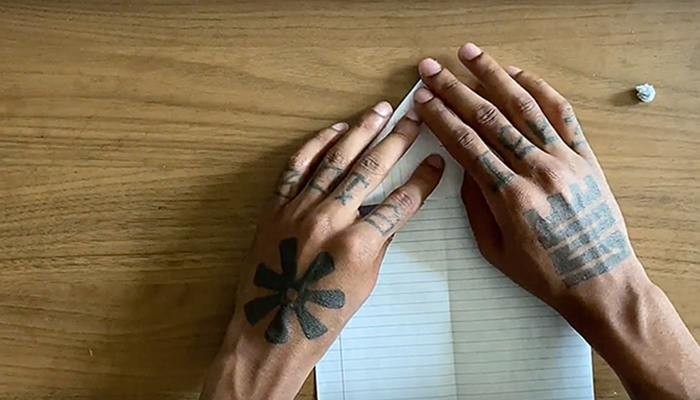 The tattooed hands of African American artist Hakim Callwood begin to fold a piece of lined paper on a wooden surface into a paper airplane