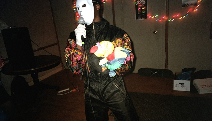 A person in a mask and jumpsuit holds a microphone in one hand and a puppet in the other.