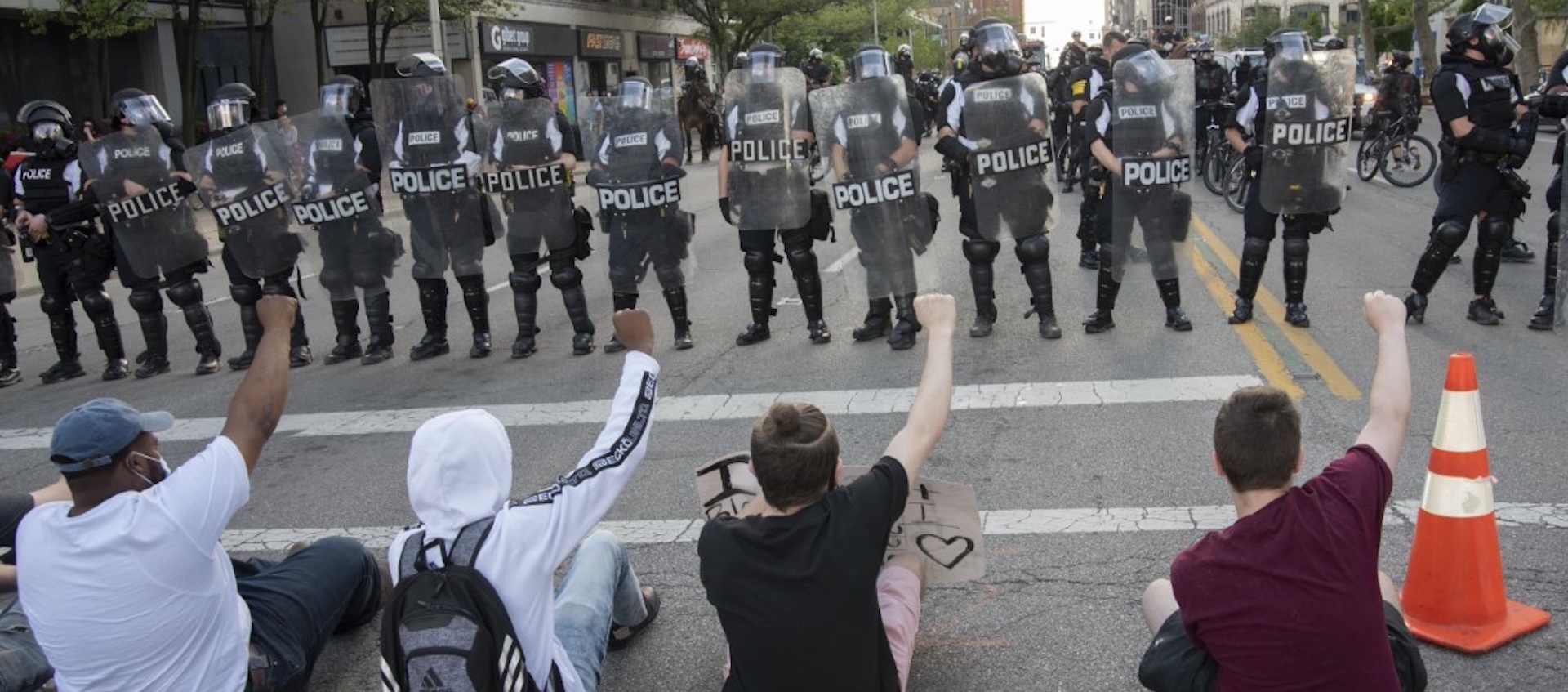 A line of four individuals protesting the death of George Floyd seated on the street, each with one fist raised, facing a line of police in riot gear