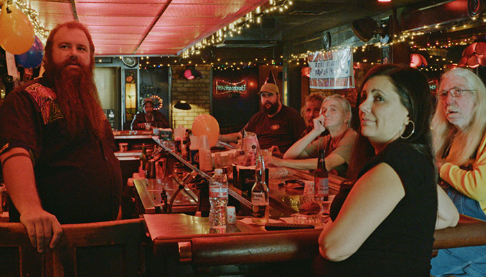 A group of six men and women of various ages sit a dive bar counter tended by a bearded man
