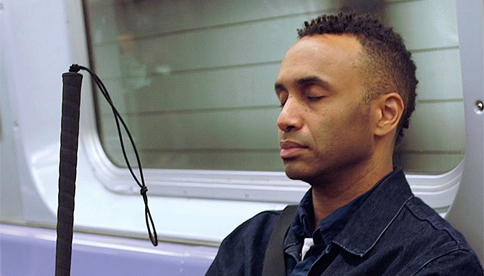 Filmmaker Rodney Evans seen shoulders up, sitting on a subway train with eyes closed, holding his cane, in a scene from his documentary Vision Portraits