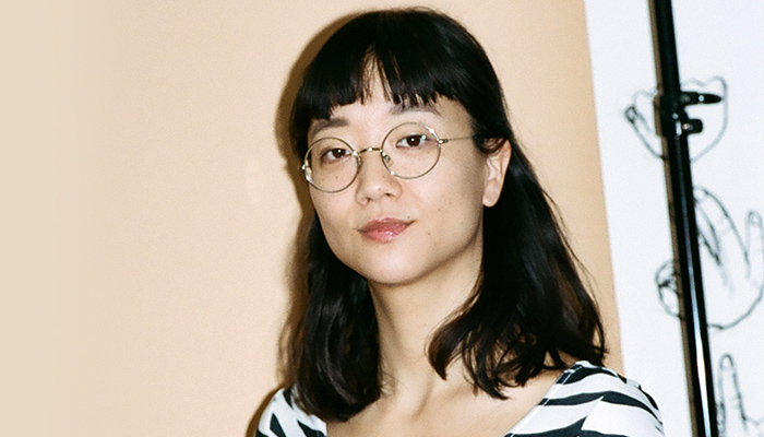 Photo of Christine Sun Kim in front of beige wall. Christine is wearing glasses and striped shirt