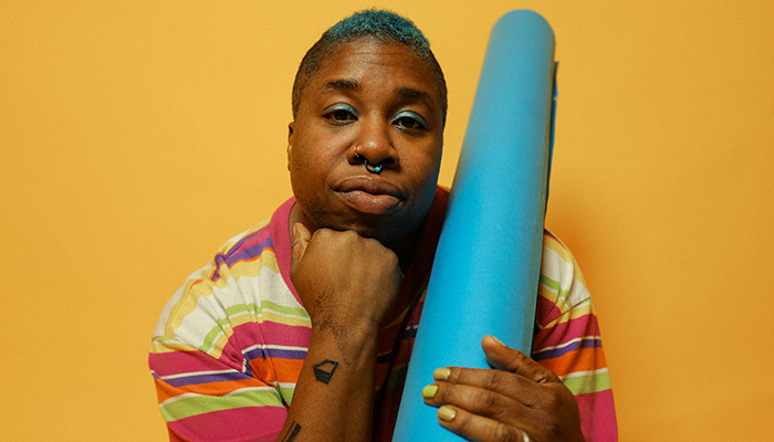 Sharon Udoh in front of a yellow wall with hand beneath chin. Sharon hold a blue tube.