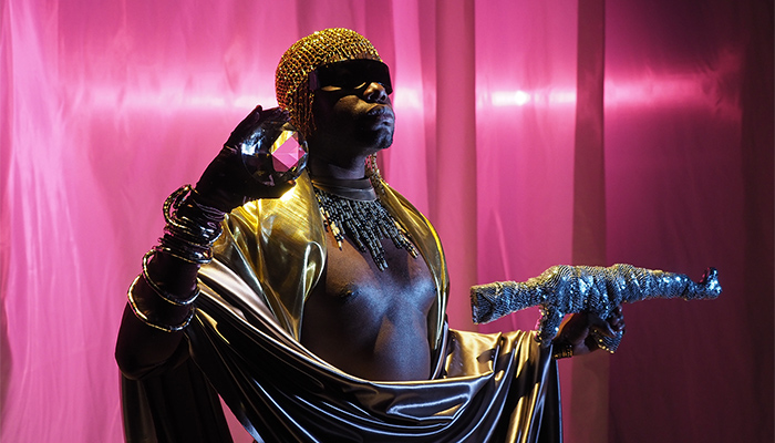 Artist Jaamil Olawale Kosoko, dressed in gold clothes and headdress, stands against a shimmering purple background while holding a crystal orb in one hand and a gun wrapped in sequined fabric.