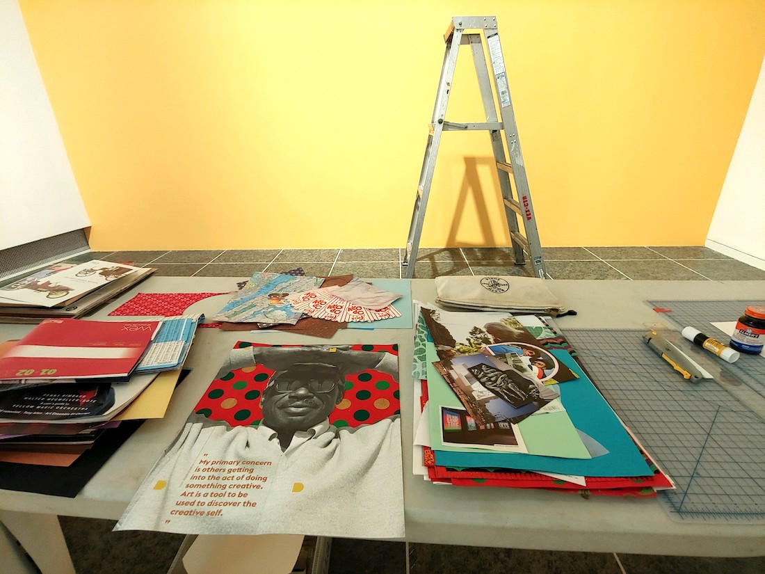 A collection of potential collage materials on a table in the foreground, with a yellow wall with a ladder in front of it in the background