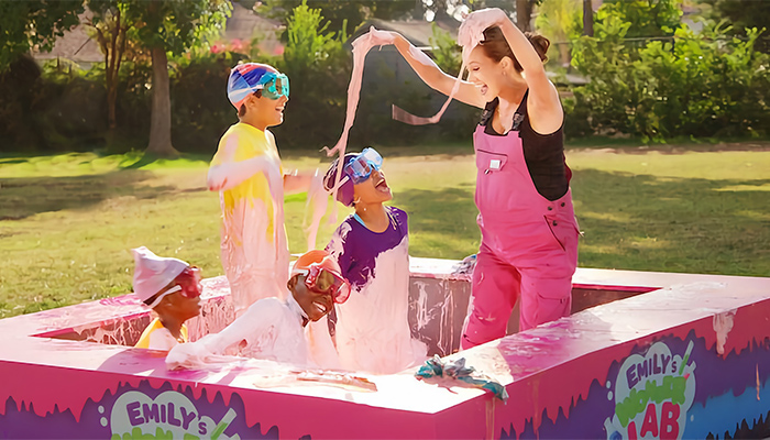 A woman in pink overalls plays with four children in a small outdoor pool filled with pink slime