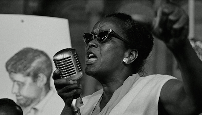 Image of Ella Baker wearing sunglasses and standing at a microphone speaking passionately