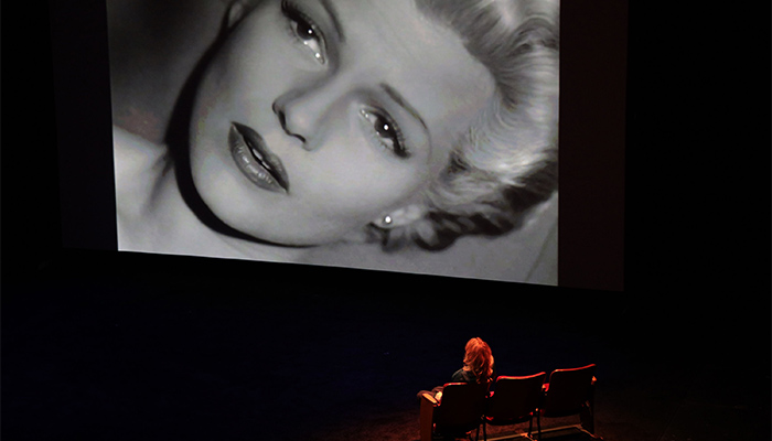 Nina Menkes sits on stage and watches a black and white film scene that depicts a woman's face up close.