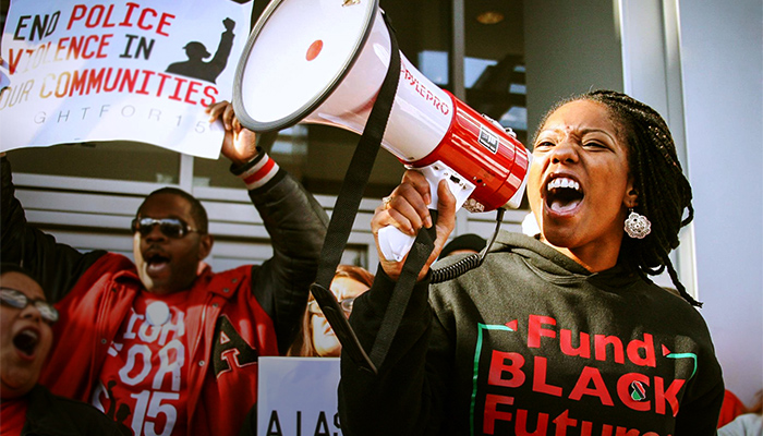 A woman shouts passionately into a megaphone while a man behind her holds up a sign supporting the end of police violence.