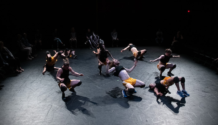A group of nine dancers wearing yellow shorts and black crop tops in a circle on a low-lit stage, all squatting or in other positions that place them close to or on the floor. An audience can be seen around them past the edge of the stage