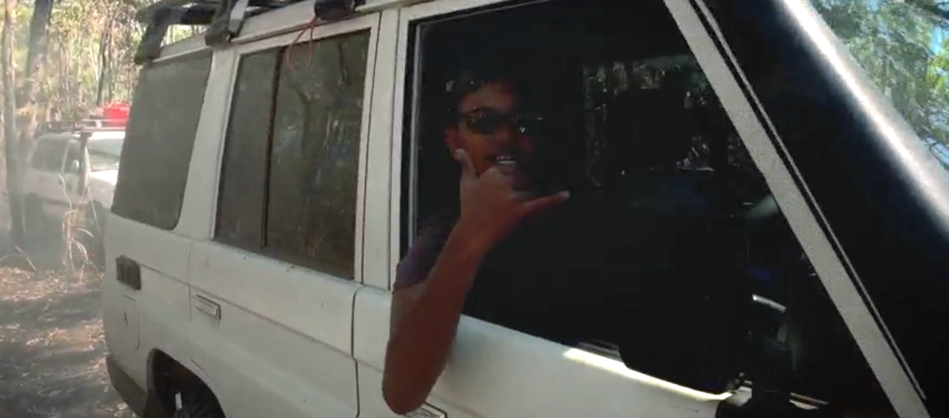 An aboriginal man leans out of the passenger side of a white SUV to make a "hang ten" hand gesture as he drives through an Australian forest. Another white SUV is driving behind him.