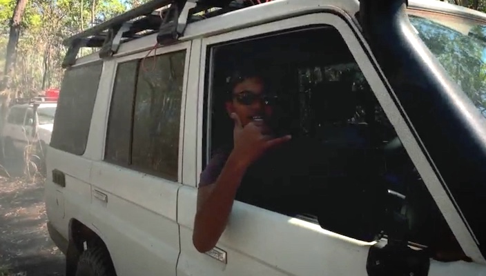 An aboriginal man leans out of the passenger side of a white SUV to make a "hang ten" hand gesture as he drives through an Australian forest. Another white SUV is driving behind him