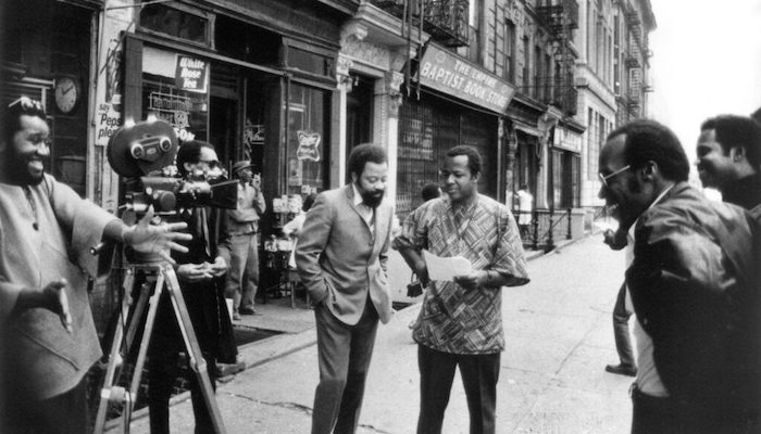 Black-and-white archive image of filmmaker William Greaves with a group of Black men shooting a film on a street full of storefronts