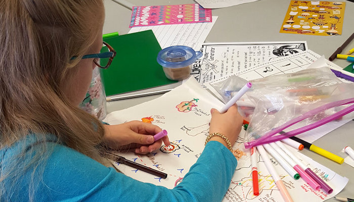 A young blonde girl in a green shirt and glasses, seen partly from behind as she draws in a sketchbook on a table covered in papers and art supplies
