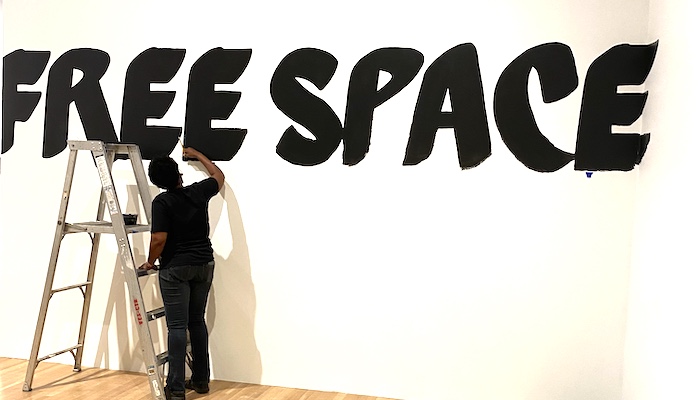 Artist Lisa McLymont is seen from behind, with a ladder beside her, paints the words "free space" in black on a white wall
