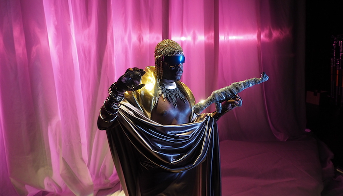 Artist Jaamil Olawale Kosoko stands on a stage with a pink fabric backdrop, wrapped in a gray silk sheet, wearing a metallic gold scarf, bejeweled crocheted beanie and gloves with bangle bracelets. In his left hand he holds what appears to be a rifle wrapped in sequined fabric 