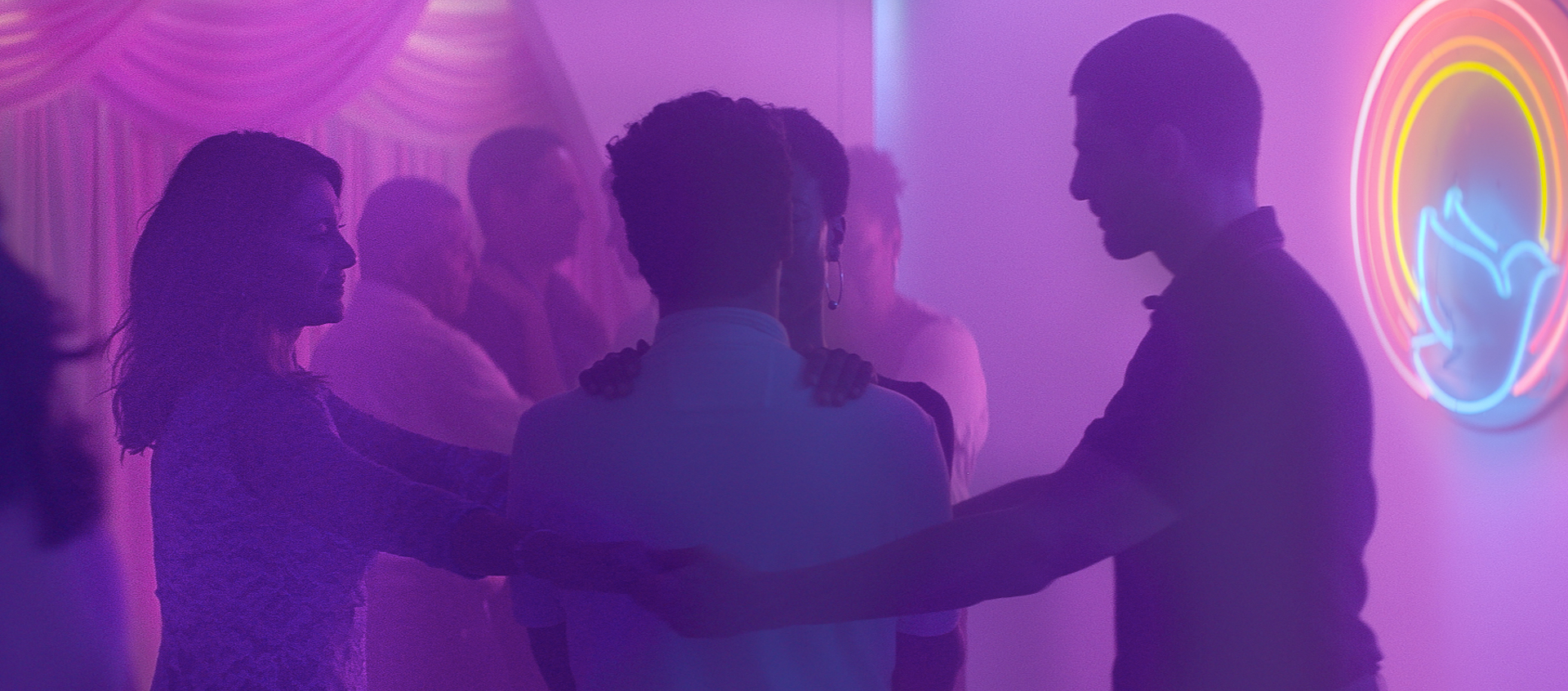 A husband and wife clasp their hands around another husband and wife couple in an embrace in a room lit by purple neon in a scene from Gabriel Mascaro's Divine Love