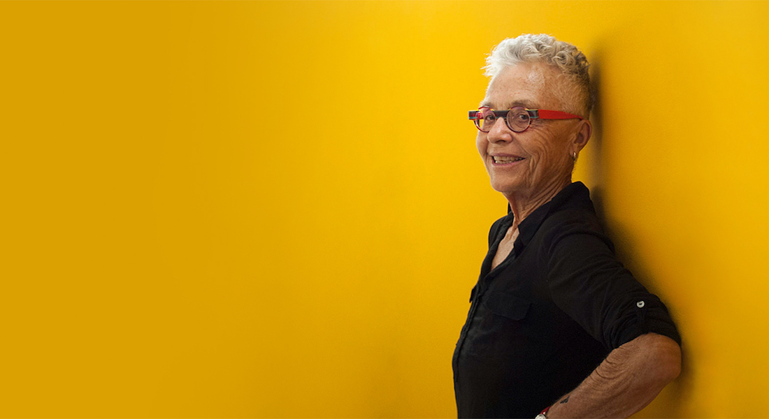 Filmmaker Barbara Hammer standing in profile against a solid yellow background, turned toward the camera and smiling