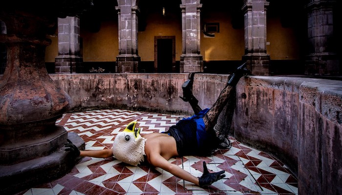 Jessica Bastidas from the group La Pocha Nostra lies face down on a tiled floor in the middle of a round space filled with aged columns, topless with a lacy skirt, tights and high heels, a large eagle mask covering her head