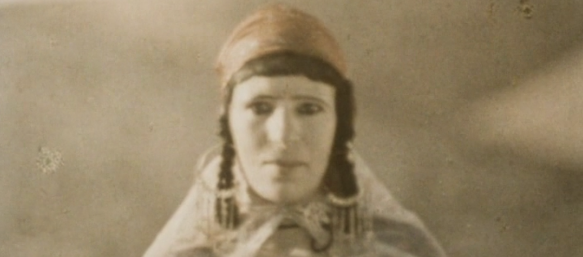 Sepia tone mage of a woman facing the camera wearing a head scarf, braids, large hoop earrings and a cape from Assia Djebar's 1982 French documentary Zerda and the Songs of Forgetting