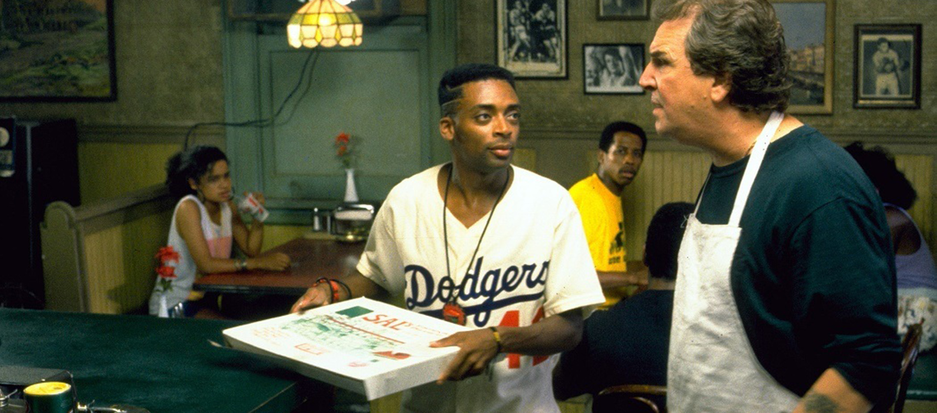 A young man in a Dodgers jersey holding a pizza box looks at an older man wearing a white apron to his right
