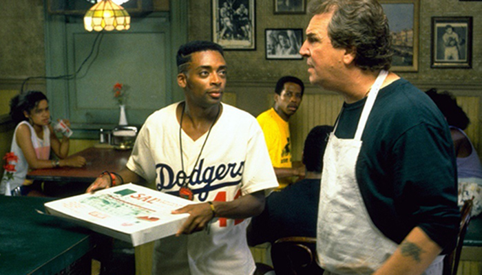 A young man in a Dodgers jersey holding a pizza box looks at an older man wearing a white apron to his right