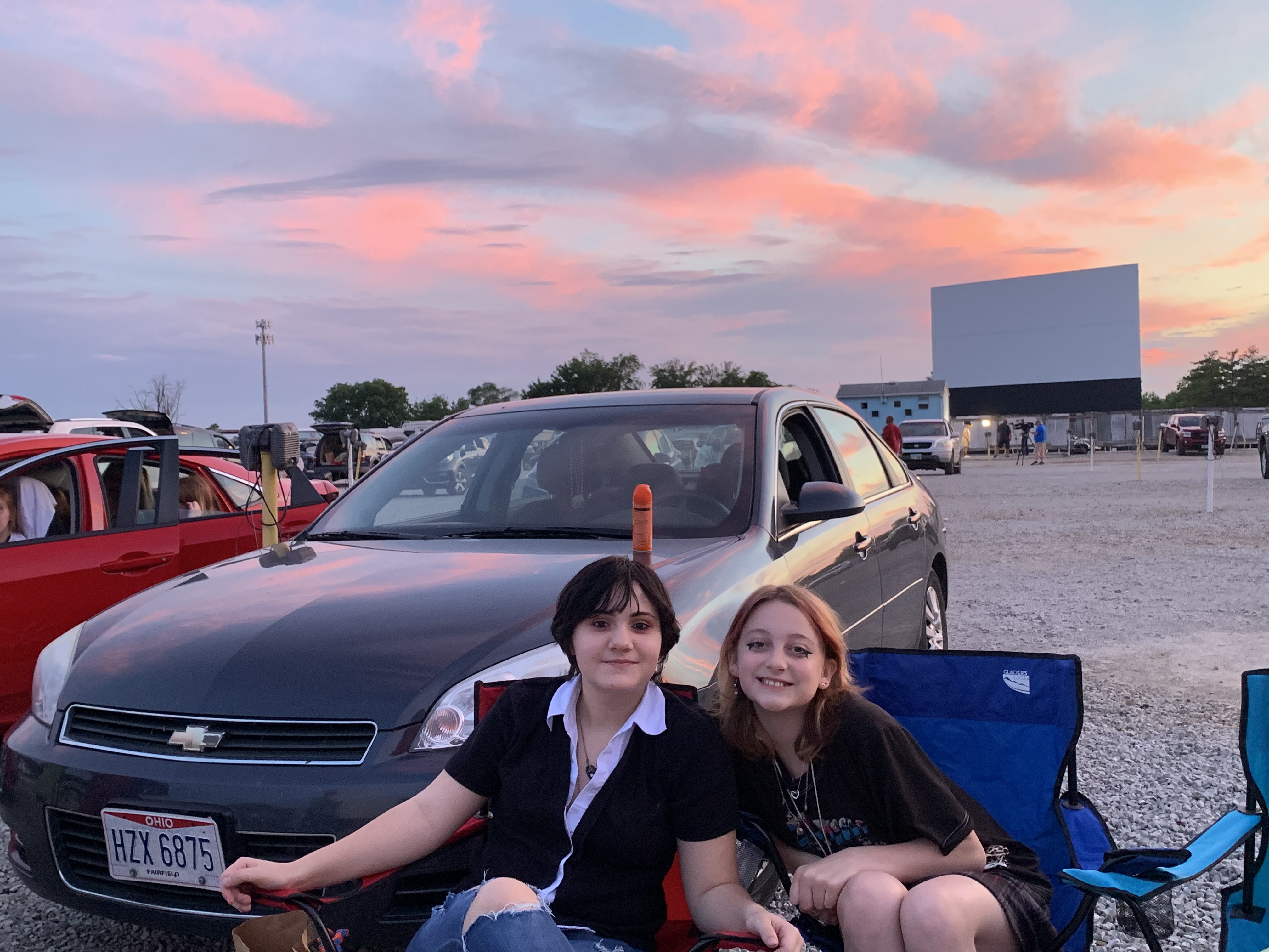Two teenage girls sit together outside a car at the drive-in