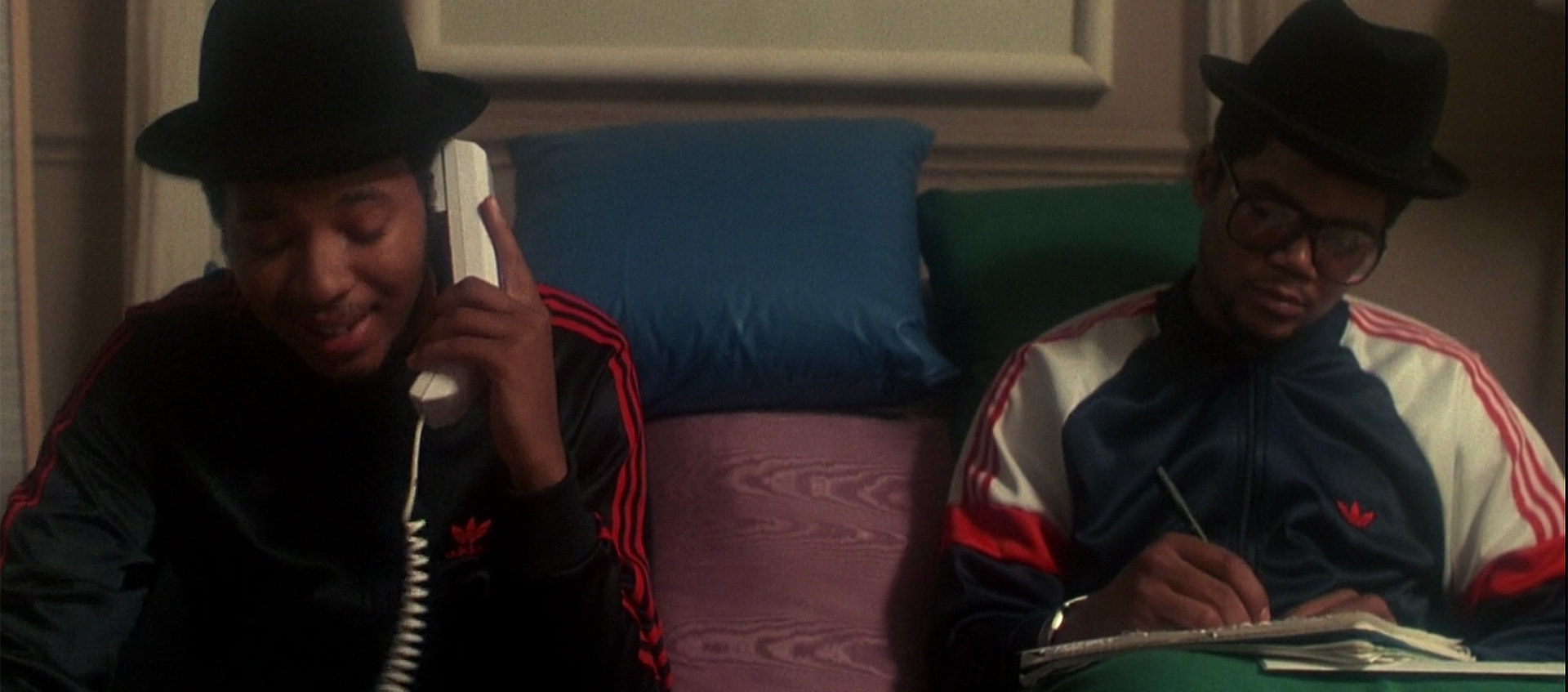 Two members of RUN DMC sit on a couch, one is on the phone while the other writes in a notebook