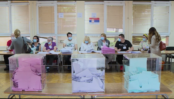 a scene of individuals checking in at a long table of volunteers to vote in the Serbian parliamentary election of June 2020. A trio of clear plastic boxes on a table, each holding ballots on paper that is pink, white, or light blue, are seen in the foreground